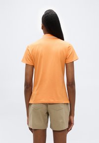 Personne aux cheveux longs portant un polo à manches courtes orange et un short beige, debout devant un fond uni clair.