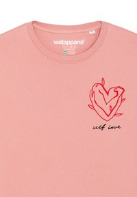 T-shirt en coton rose clair avec un graphisme de cœur rouge entouré de feuilles et le texte "amour de soi" en dessous. Encolure ras de cou et texture douce.