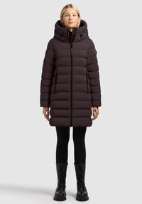 Lila Steppjacke mit hohem Kragen und großer Kapuze, ausgestattet mit einem Frontreißverschluss, seitlichen Taschen und gerippten Bündchen. Getragen über schwarzen Leggings und Stiefeln.