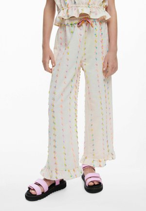 Dieťa oblečené v bielych nohaviciach s viacfarebnými zvislými bodkami a volánikmi na lemoch, v kombinácii s ružovými sandálmi na bielom pozadí.