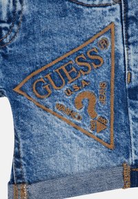 Pantalones cortos de mezclilla en azul claro con un efecto desgastado. Presenta un parche triangular bordado marrón con "GUESS U.S.A" y un diseño de signo de interrogación.