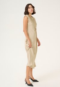 Abito beige senza maniche con dettaglio annodato in vita, tessuto fluido e lunghezza midi. Décolleté nere con tacco a schiocco e accentatura a fiocco.