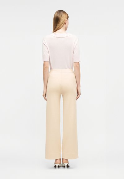 Γυναίκα που στέκεται με την πλάτη γυρισμένη, φοράει ροζ ανοιχτό ροζ κοντομάνικη μπλούζα, μπεζ παντελόνι με φαρδιά μπατζάκια και άσπρα παπούτσια με τακούνι σε απλό φόντο.