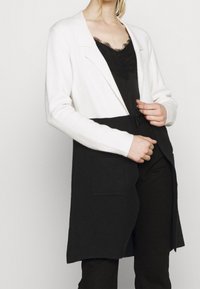 Long cardigan featuring a white top half and black bottom half, with a lapel collar and two patch pockets on the front. Soft texture.