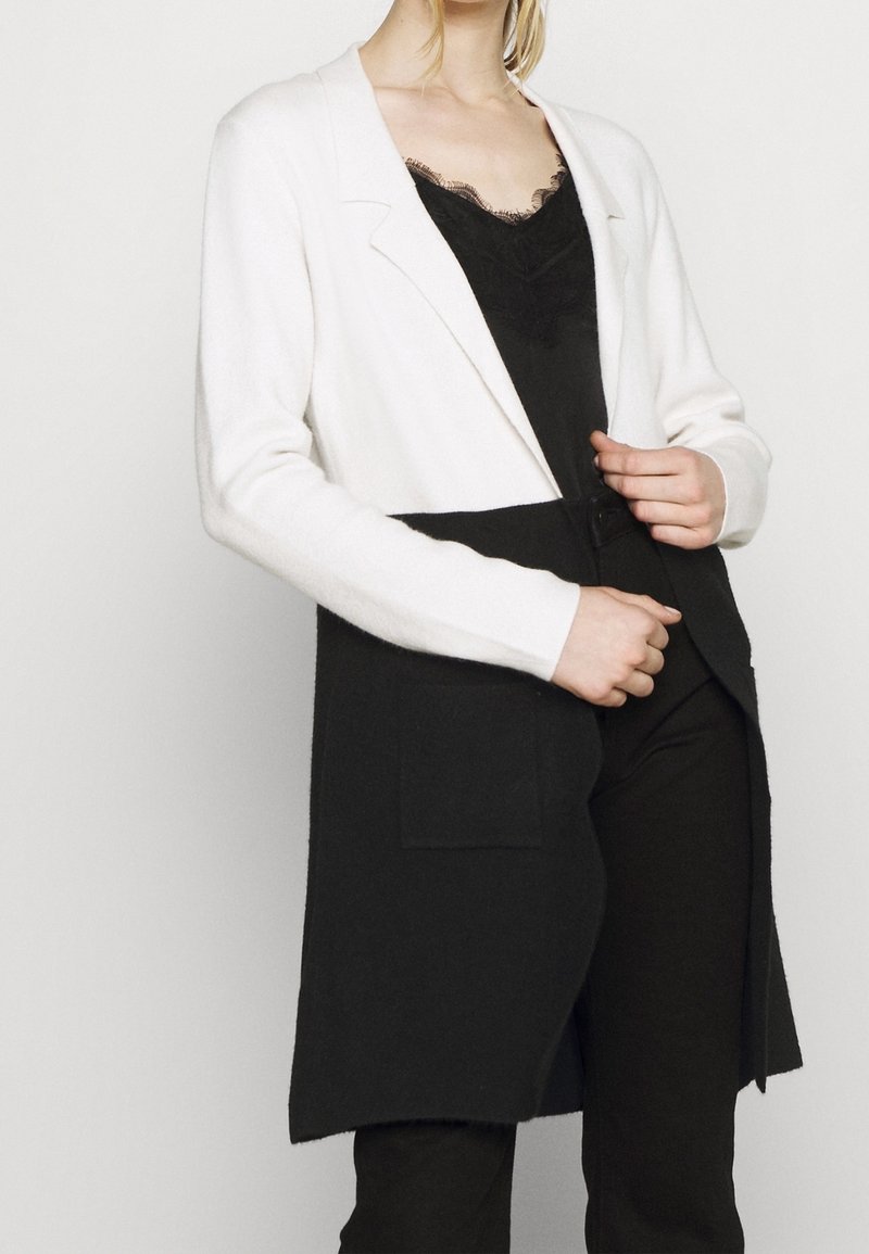Long cardigan featuring a white top half and black bottom half, with a lapel collar and two patch pockets on the front. Soft texture.