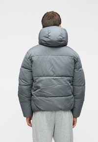 Inimene seljaga pöördunud, kandes halli sulejope, millel on kõrge polsterdatud krae ja heledad hallid dressipüksid, valge taustaga.