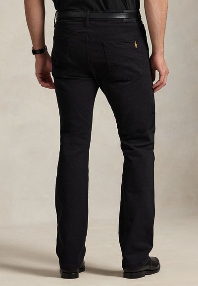 Man wearing black straight-leg pants with a black belt and black shoes, standing against a plain beige background.