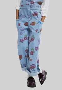 Salopette in denim caratterizzata da un tessuto azzurro chiaro con fantasie di frutta e farfalle colorate, con bordi arrotolati e tasche laterali.