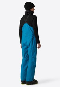 Blauw waterdicht overall met verstelbare banden, voorzien van een zwarte achterkant en praktische zakken. Draag het over een zwart longsleeve shirt en beanie.