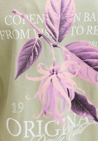 Vaaleanvihreä t-paita, jossa on suuri violetti kukkakuosi, yksityiskohtaiset terälehdet ja lehdet, ja mukana valkoisia tekstielementtejä, kuten "ORIGINAL".