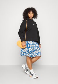 Sweatshirt preto com capuz, saia floral azul e branca, mala de palha amarela e ténis brancos com realces em cinza e laranja.