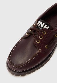 Zapato de barco de cuero marrón con costuras blancas y ojales dorados. Presenta una puntera redonda y una etiqueta de marca en blanco y negro en la lengüeta.