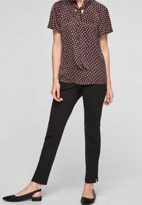 Dunkelbraune Bluse mit einem Halsband, die ein sich wiederholendes Augenmuster in hellrosa und weiß aufweist. Getragen mit engen schwarzen Hosen und Slingback-Ballerinas.