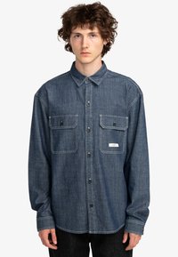 Mørk denim skjorte med knapper, to brystlommer, krave og kontraststing. Lange ærmer og almindelig pasform, vist på en model.