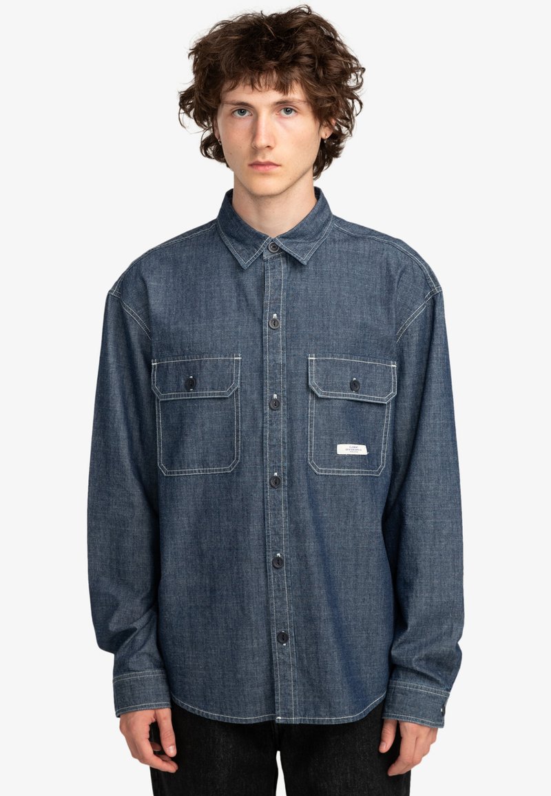 Mørk denim skjorte med knapper, to brystlommer, krave og kontraststing. Lange ærmer og almindelig pasform, vist på en model.
