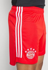 Pantaloni sportivi rossi con tre strisce bianche lungo il lato, con logo ricamato dell'FC Bayern München e cinque stelle sopra di esso.