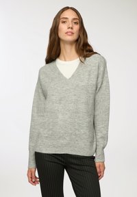 Maglione v-neck grigio chiaro con maniche lunghe, realizzato in un tessuto morbido e testurizzato, abbinato a pantaloni neri a righe sottili. Design semplice e moderno.