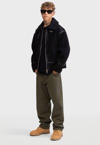 Veste en polaire noire avec fermeture éclair et col, accompagnée d'un t-shirt noir, d'un pantalon vert olive et de bottes de travail beiges. Les accessoires comprennent des lunettes de soleil noires.