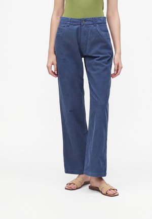 Personne portant un pantalon en velours côtelé bleu droit, un haut vert et des sandales plates beiges à bout ouvert, debout sur fond blanc.