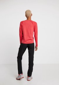 Červená košile s dlouhým rukávem a texturovanými rameny, spárovaná s černými kalhotami a šedými teniskami, viděná ze zad na jednobarevném pozadí.