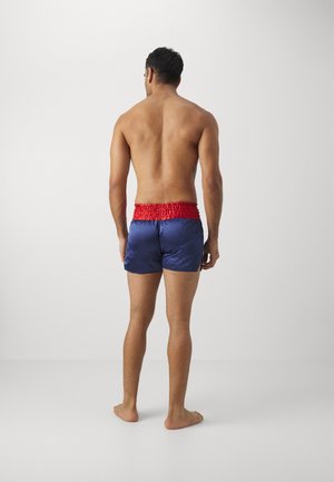 Skjorteløs mand stående barfodet, vendt væk, iført marineblå shorts med bred rødt elastikbånd mod en ensfarvet hvid baggrund.