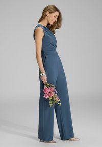 Blauwe jumpsuit met gestructureerde schouders, getailleerde taille, wijde pijpen, gecombineerd met zilveren sieraden en een boeket rozen bloemen.