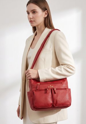 Woman wearing cream blazer carries red B Cavalli leather shoulder bag with multiple zippered pockets against plain background.