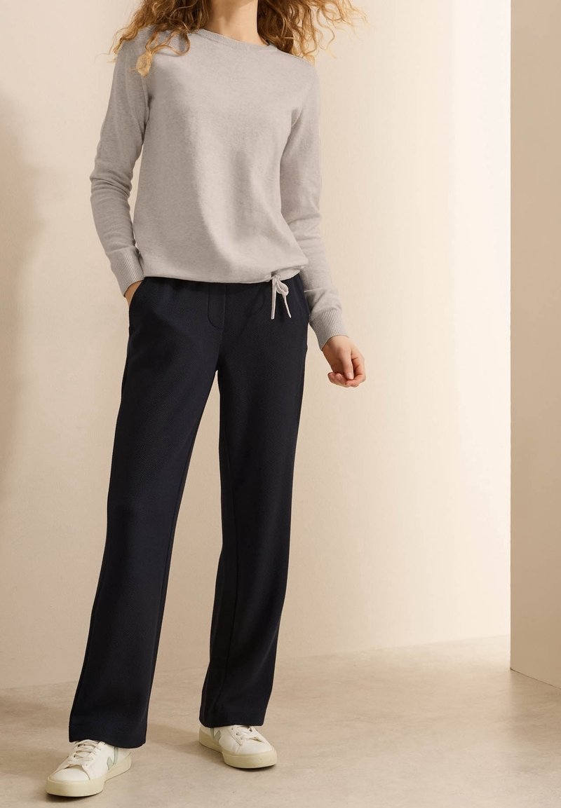 Une personne porte un pull à manches longues gris clair et un pantalon noir ample avec des baskets blanches, debout sur un sol clair contre un mur uni.