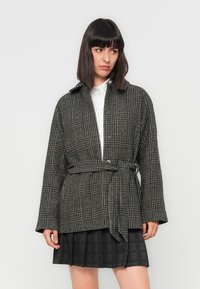Black and white checkered coat with a belted waist, featuring a collar and button-down front. Textured fabric, loose fit, and long sleeves.