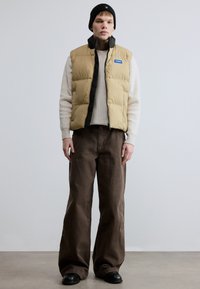 Beež puffer vest musta voodriga, tõmbluku kinnitusega ja sinise logoga, kombineeritud valge soonikkoega kampsuni ja pruunide laia säärega pükstega. Must talvemüts.