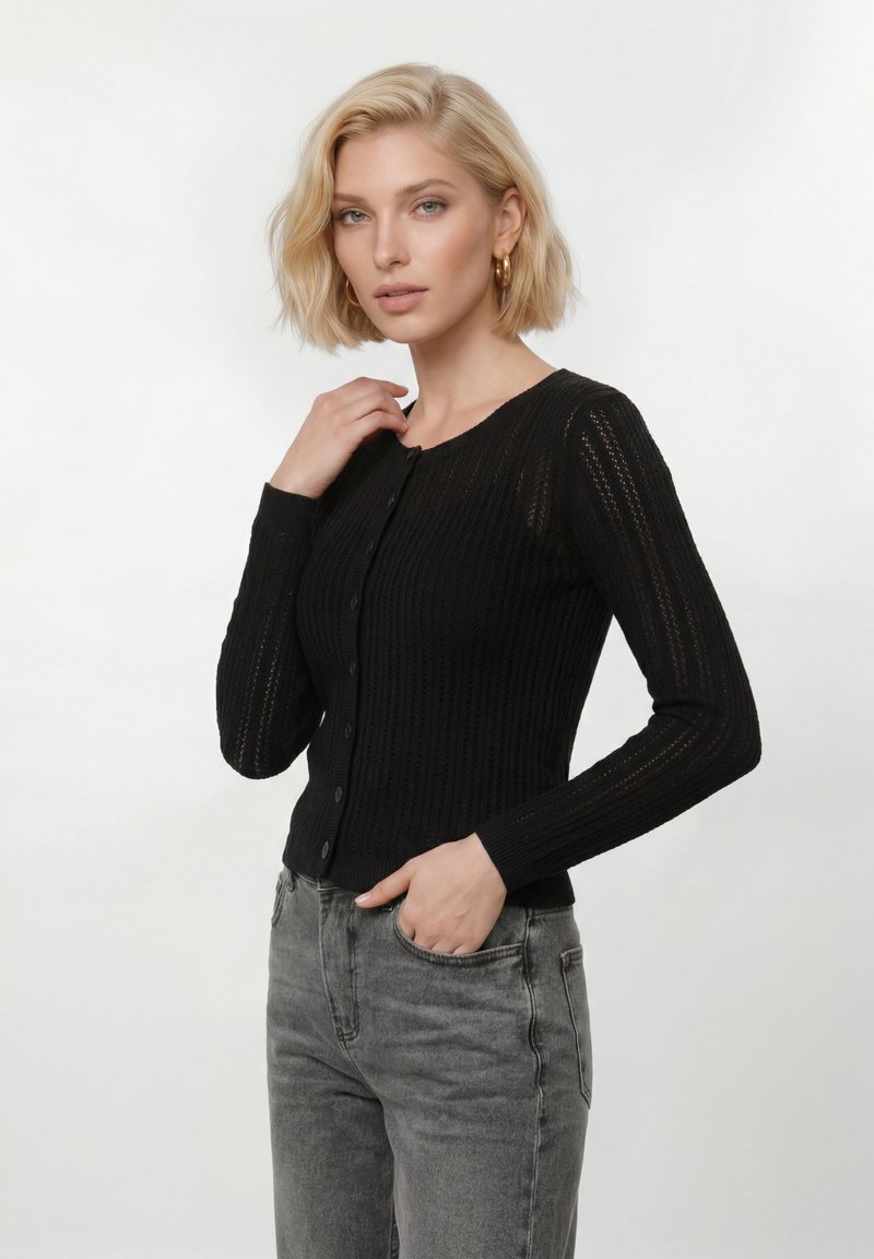 Blonde Frau, die goldene Creolen, einen schwarzen, geknöpften Strickcardigan und graue Jeans trägt, posiert mit einer Hand am Kragen und der anderen in der Tasche.