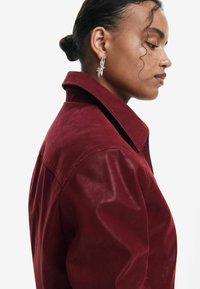 Veste en cuir bordeaux avec un col structuré et des coutures visibles. Le modèle porte des boucles d'oreilles en argent en forme d'étoile ; sa coiffure est un chignon bas.