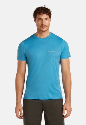 Hombre con cabello rizado que lleva una camiseta azul de manga corta con cuello redondo y el logo de "icebreaker", y pantalones oscuros, sobre un fondo blanco.