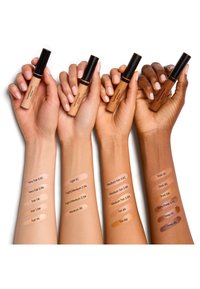 Vier armen met diverse huidtinten, elk houdend een tube concealer; streeptestjes met namen op elke arm tonen verschillende tinten en ondertonen.