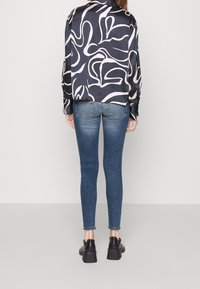 Camisa de manga larga en satén azul oscuro con patrones abstractos blancos, combinada con jeans ajustados azules y zapatos negros gruesos con tacón.