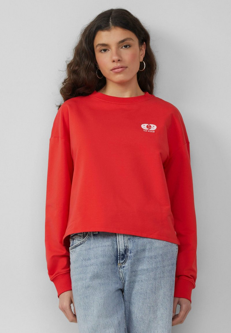 Rode sweatshirt met lange mouwen en een ronde hals, met een witte grafische afbeelding met de tekst "wees vriendelijk" op de borst. Casual pasvorm.