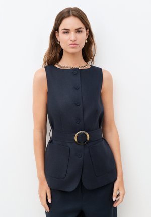 Mujer que lleva un chaleco negro sin mangas abotonado con cinturón con hebilla redonda, combinado con pantalones a juego y un collar de cadena dorada.