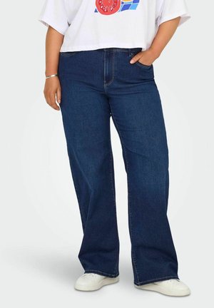 Osoba nosząca ciemnoniebieskie dżinsy z szerokimi nogawkami, białe trampki i białą krótką koszulkę z kolorowym nadrukiem, ręka w kieszeni.