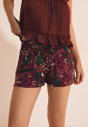 Femme portant un haut bordeaux à volants et un short à motif floral avec des accents verts et violets, la main ajustant la ceinture du short.