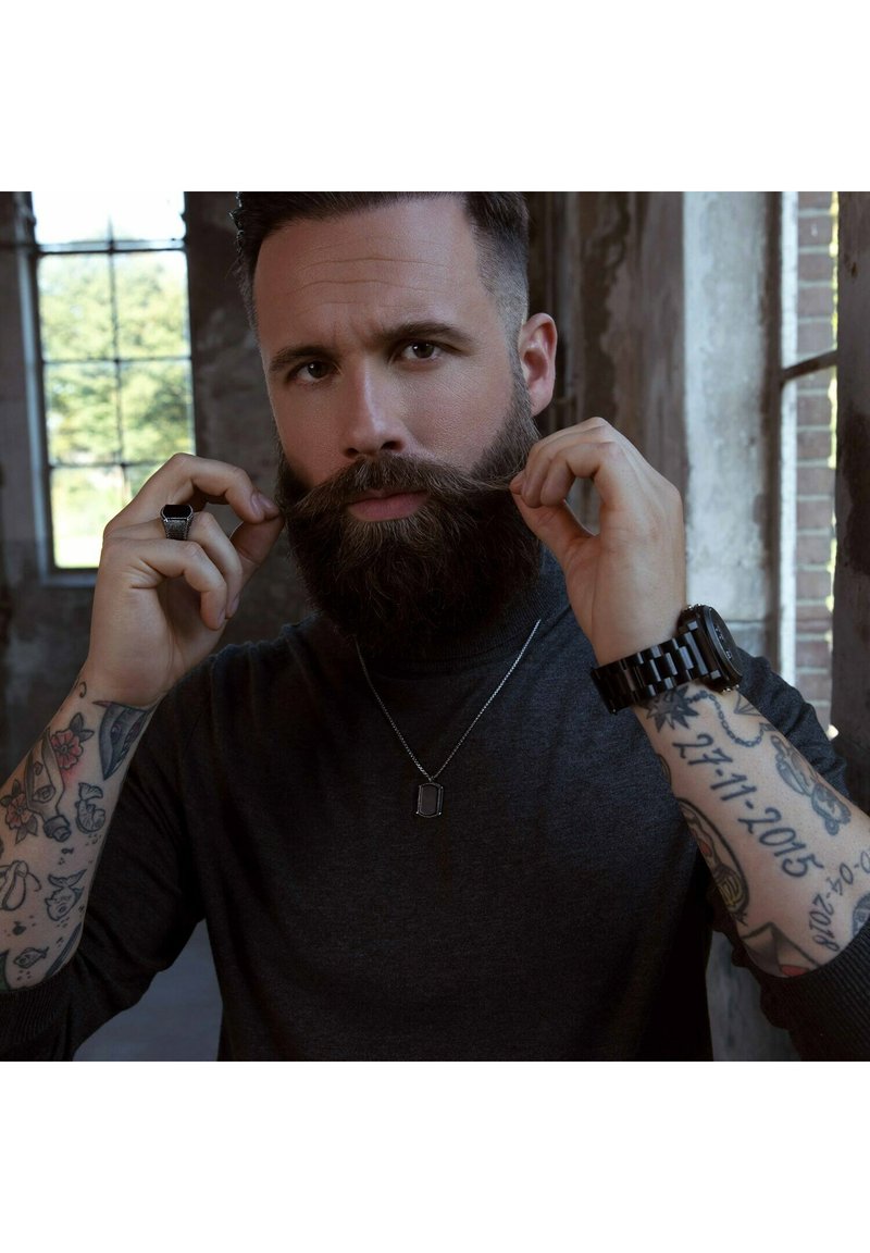 Behaarde man met kort haar, gekleed in een donkergrijze trui. Hij houdt een snor vast en toont gedetailleerde tatoeages en een zilveren ketting met een hanger.
