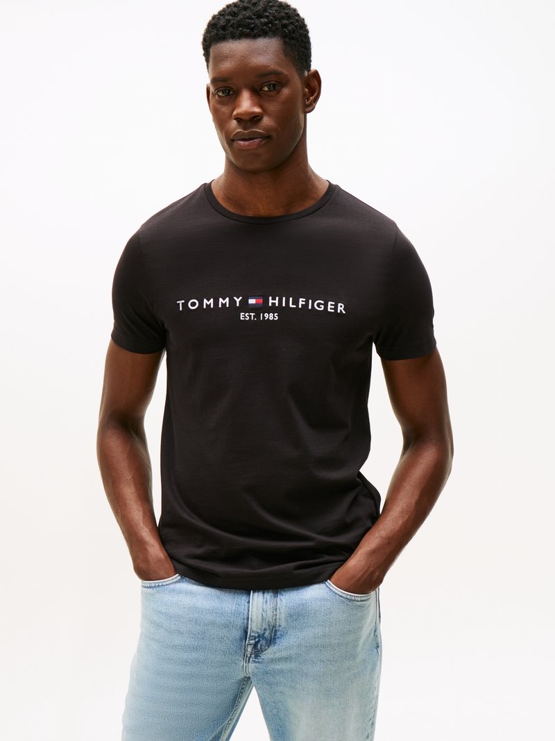 Μαύρο βαμβακερό t-shirt με λογότυπο "TOMMY HILFIGER EST. 1985" σε λευκό. Το μοντέλο φοράει ανοιχτό μπλε τζιν παντελόνι. Απλός, κλασικός σχεδιασμός.