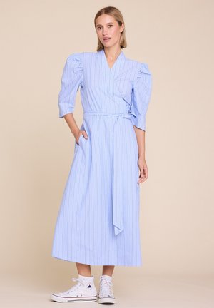 Kvinne iført en lys blåstripet midikjole med puffede 3/4-ermer, knyttet midjebelte og hvite høye sneakers, stående mot en beige bakgrunn.