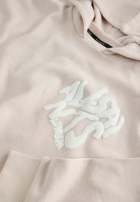 Světle růžová mikina s kapucí s texturovaným bílým grafickým designem na přední straně. Má klokaní kapsu a ribbované manžety. Vyrobena z měkké tkaniny.