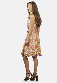 Vestido de manga corta en color naranja brillante con patrones florales y geométricos, que cuenta con un cinturón anudado y un dobladillo multicolor. Combinado con sandalias de plataforma de tacón alto marrones.