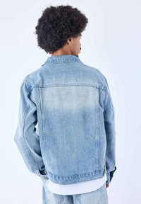 Giacca in denim blu chiaro con un design classico, caratterizzata da un colletto, tasche laterali e una texture leggermente sbiadita. Vista posteriore mostrata.