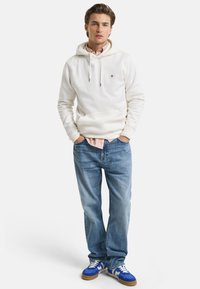 Hvid hættetrøje med lomme foran, parret med lyseblå jeans og blå sneakers. Outfittet har en lagdelt stribet skjorte under.