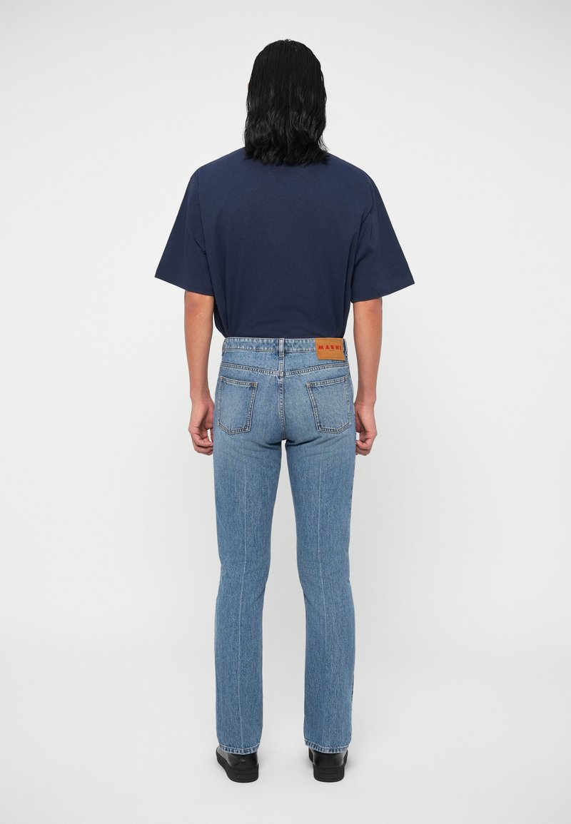Person mit schulterlangem, dunklem Haar, der ein navyblaues T-Shirt, hellblaue geradlinige Jeans und schwarze Schuhe trägt und mit dem Rücken steht.