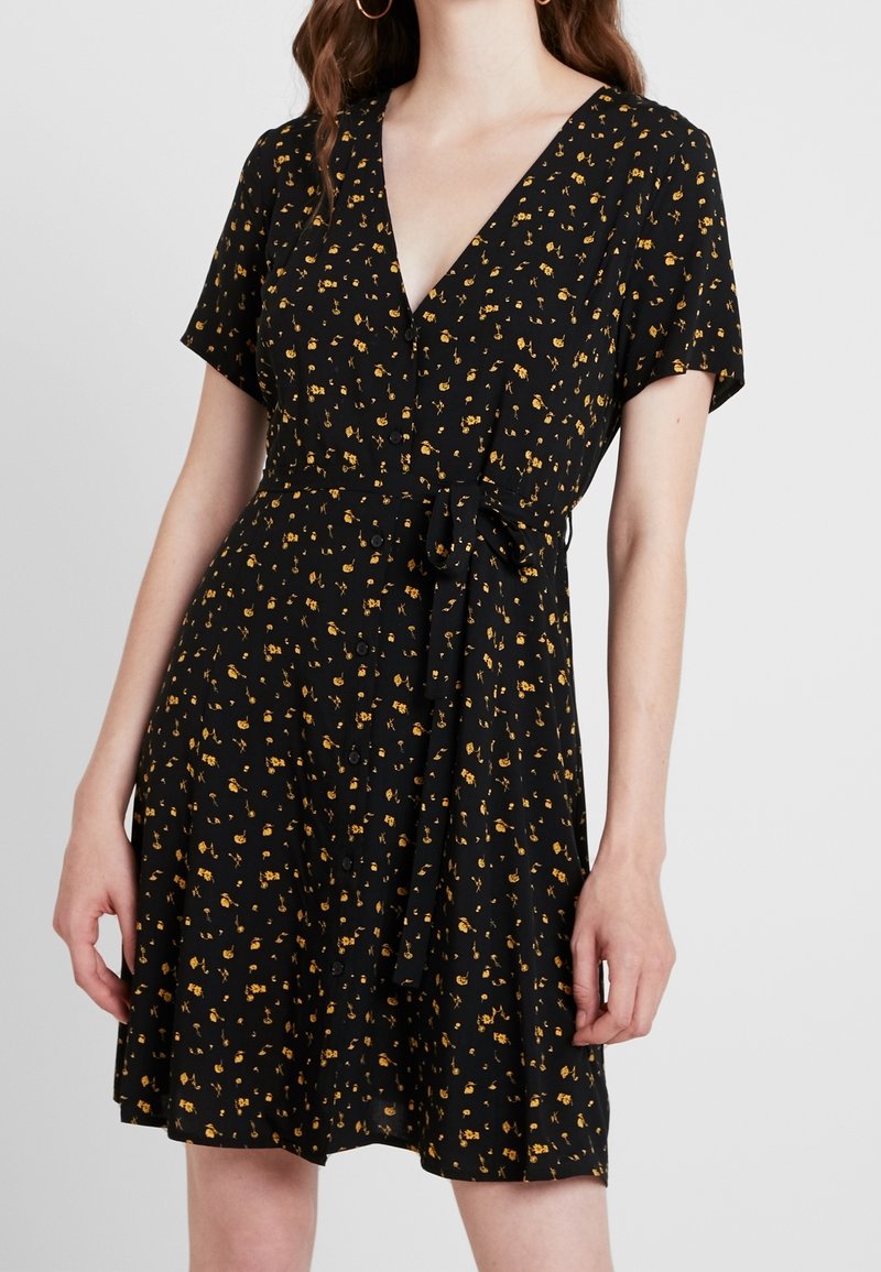 Femme portant une robe noire à manches courtes avec un petit imprimé floral jaune, col en V, boutons à l'avant et ceinture nouée à la taille, debout devant un fond uni.