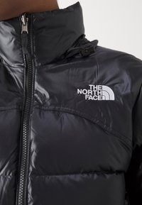 Črna izolirana jakna iz sijočega, quilted materiala z visokim ovratnikom in zadrgo. Ima bel logo na prsih.