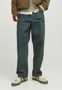 Tumed sinised denim püksid lõdva lõikega, mis sisaldavad nähtavat õmblust ja kergelt kulunud tekstuuri. Kombineeritud beežide tossudega.